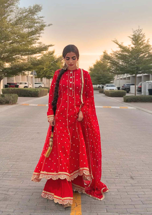 Woman in a red Pakistani and Indian  traditional outfit standing on a street with trees and vehicles in the background.
